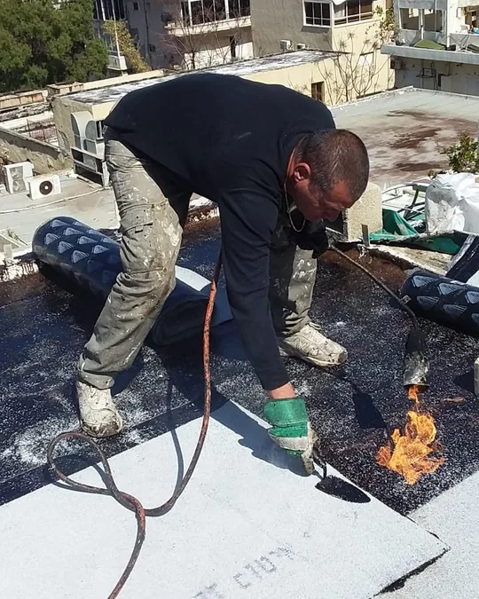 פועל מלחים יריעת איטום עם מבער על גג שטוח, הלהבה נראית והיריעה הלבנה נצמדת לתשתית